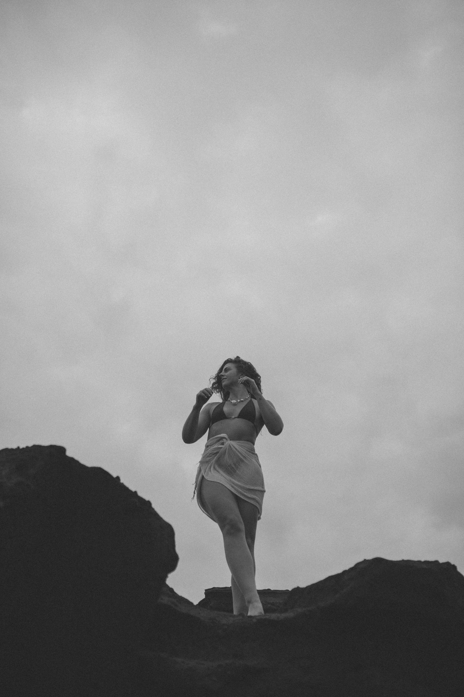 Black and white photo of woman in bikini standing on dark rocks against cloudy sky in New Zealand.