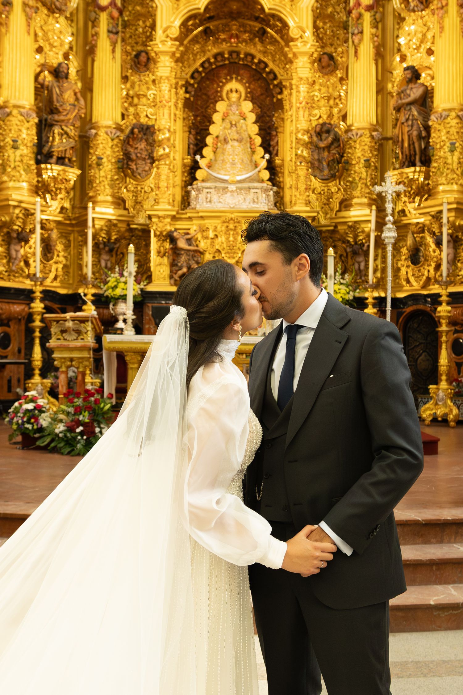 A wedding couple shares a romantic moment in an ornate golden Catholic church altar with baroque decorations.