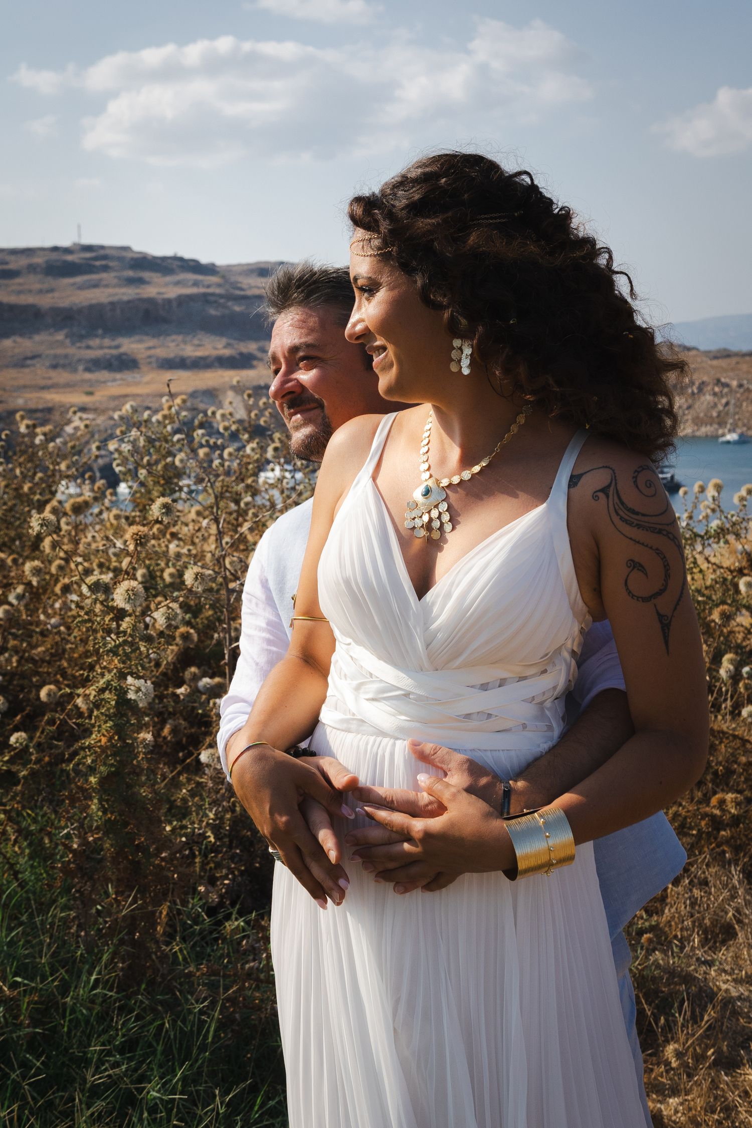 A romantic couple in white dresses embrace while standing near a coastal landscape with golden vegetation and blue waters.