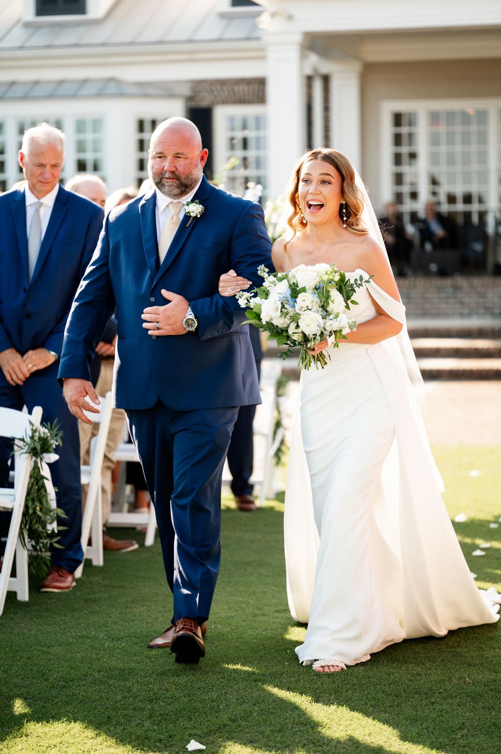 Wedding party laughs together on a sunny lawn with the bride in white and groomsmen in navy blue suits.