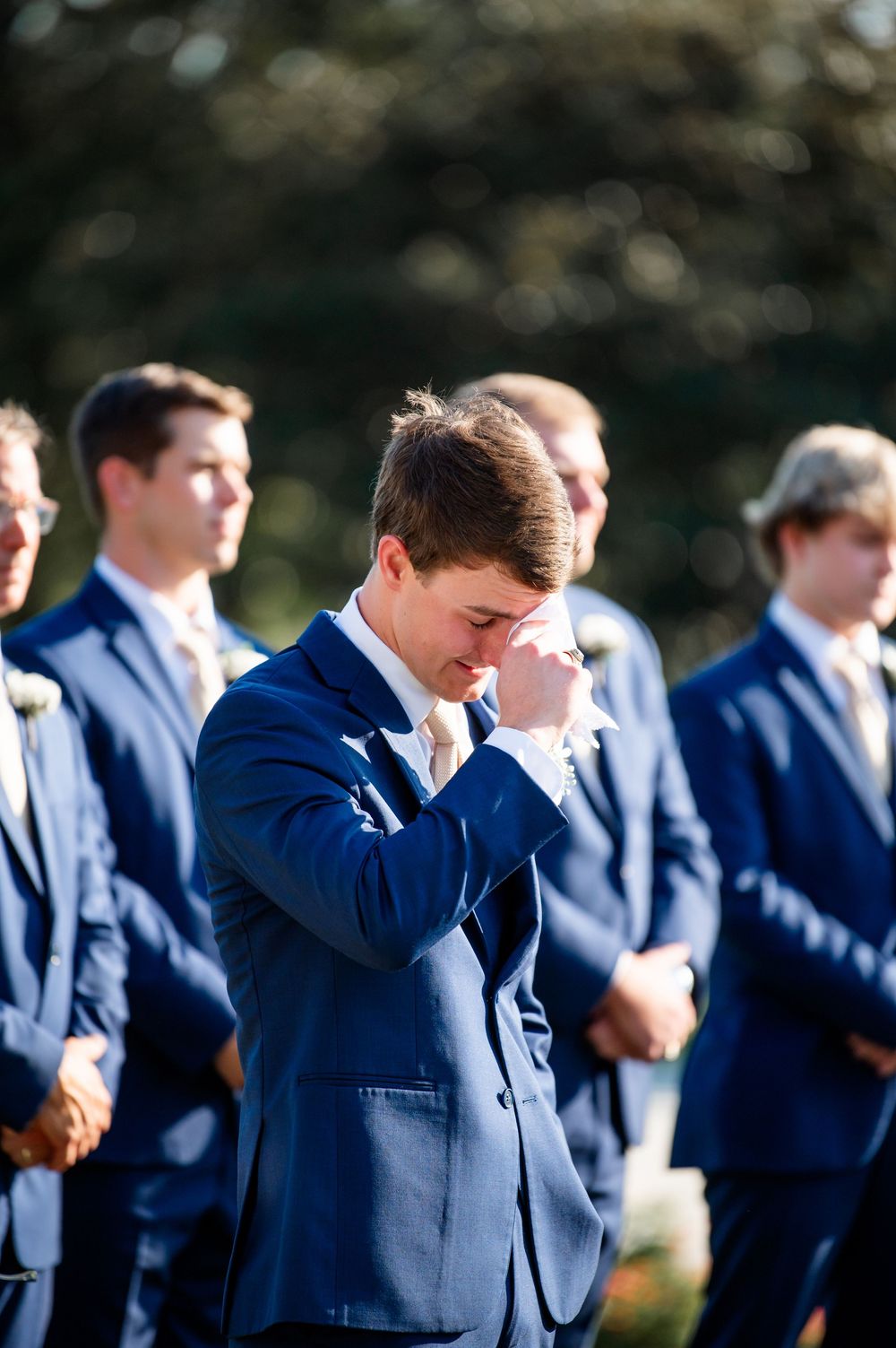 Groomsmen in navy suits bow their heads during an emotional wedding ceremony moment.