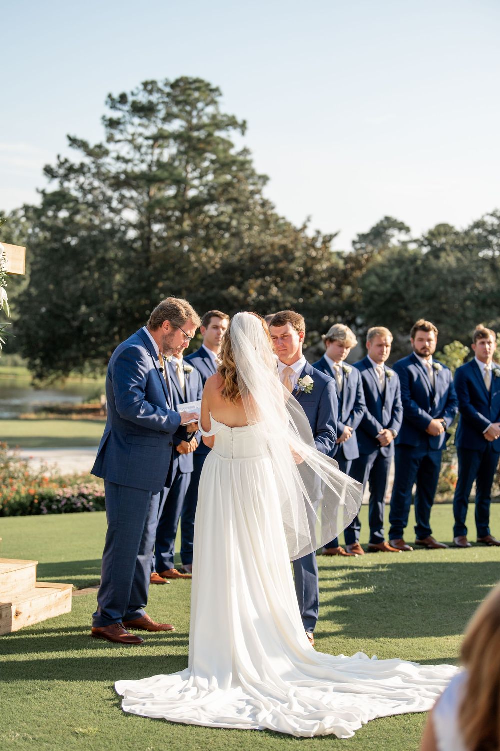 Outdoor wedding ceremony with bridal party in navy suits lined up on manicured lawn.