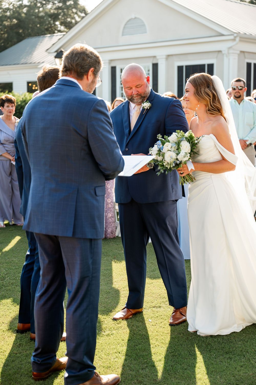 Wedding party gathered on a lawn outside a white colonial-style building.