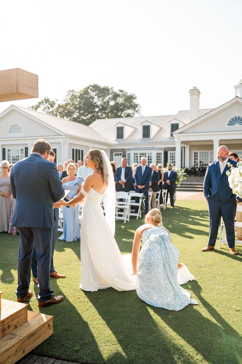 Wedding guests and party members mingle on a sunny lawn outside an elegant white venue.