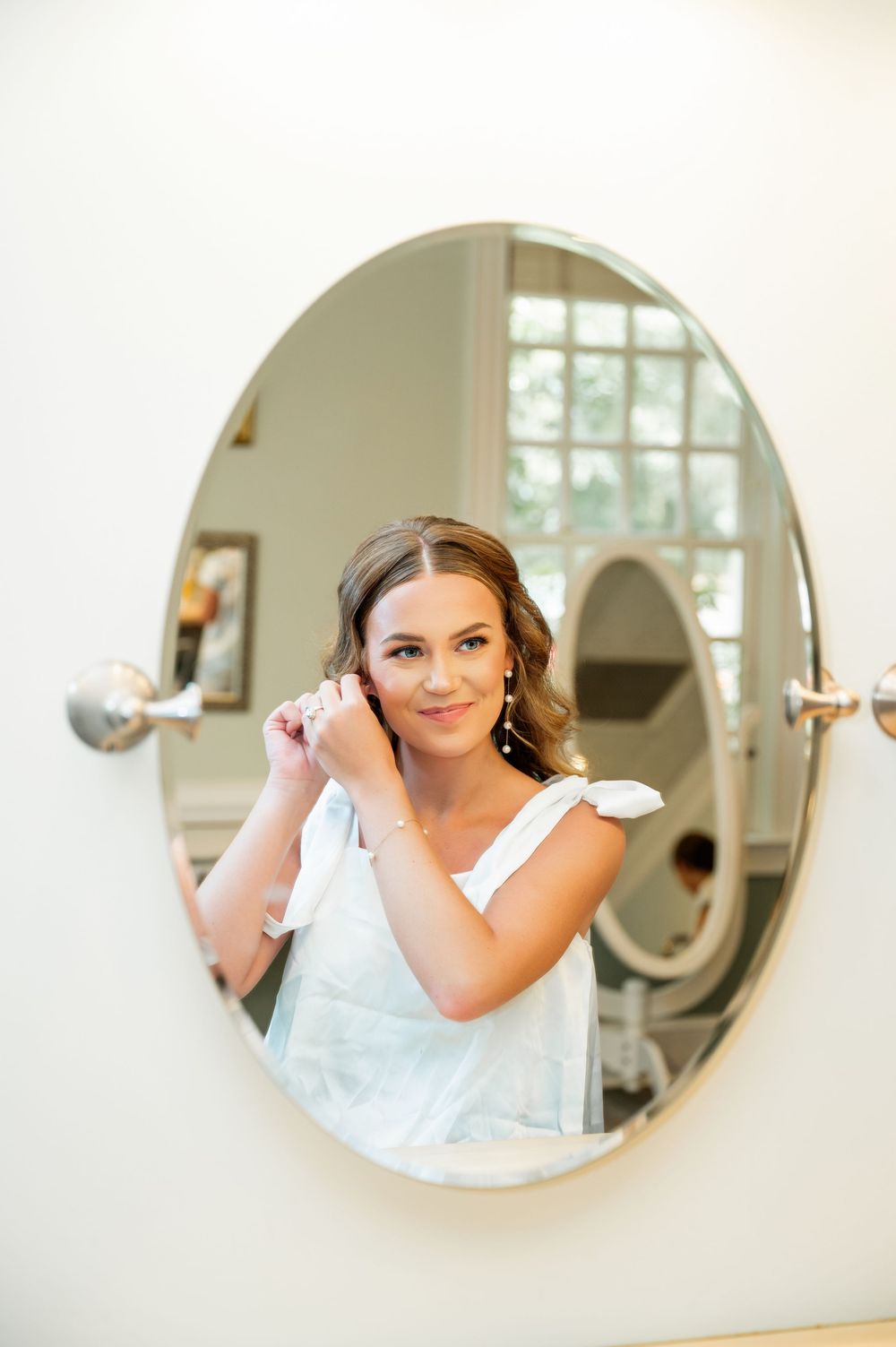 A reflection in an oval mirror shows a bride in a white dress getting ready for her wedding day.