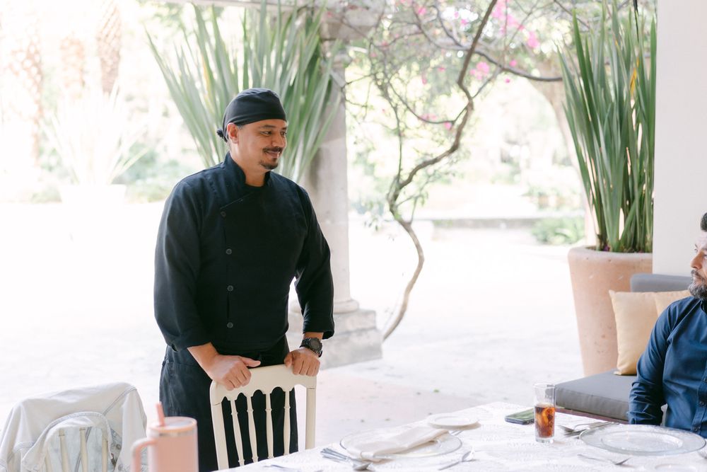 Someone in a black sweater and hat stands at a dining table with tall plants visible in the background.