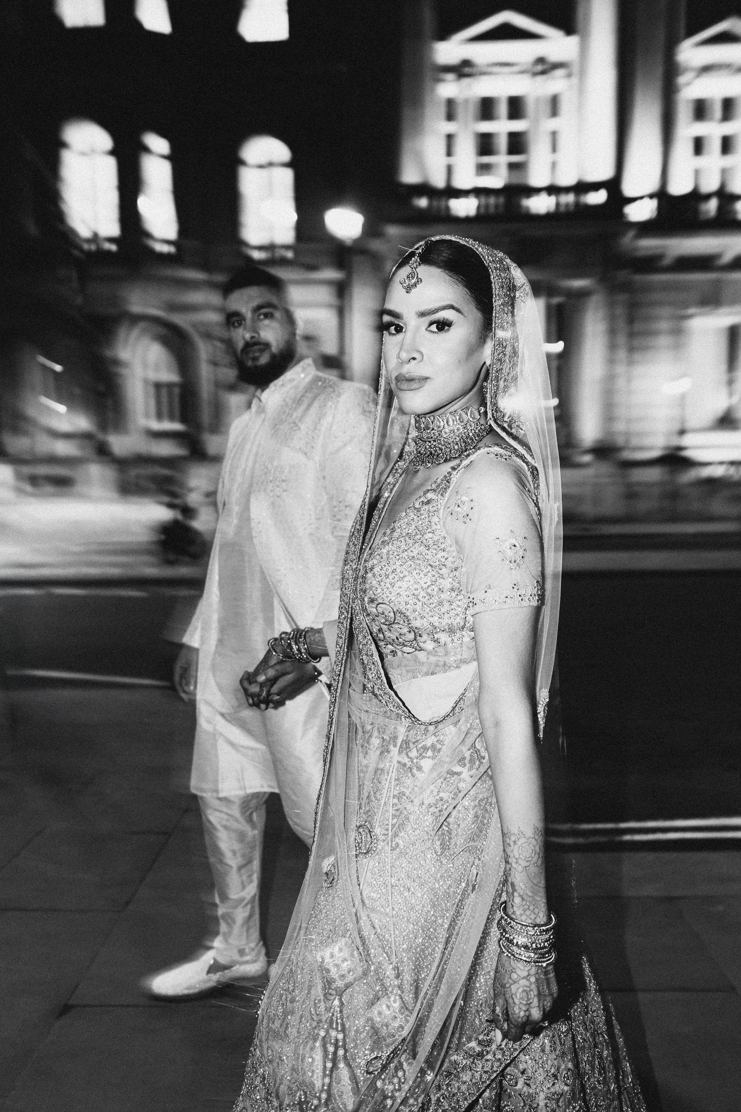 Cinematic black and white night portrait of a South Asian couple walking together outside a grand London wedding venue