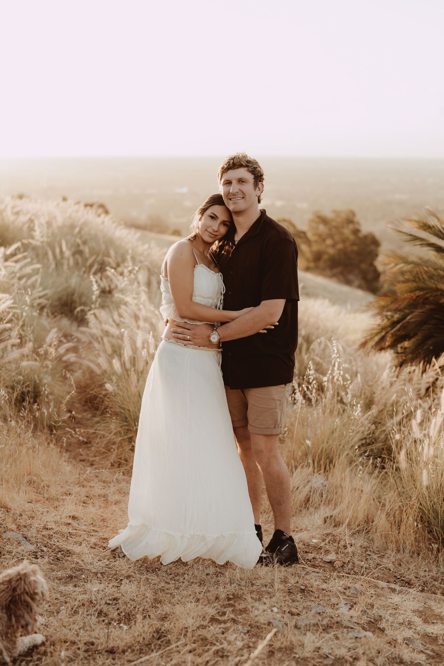 A couple embraces on a hilltop during sunset in sepia tones, surrounded by tall dried grasses and natural scenery.