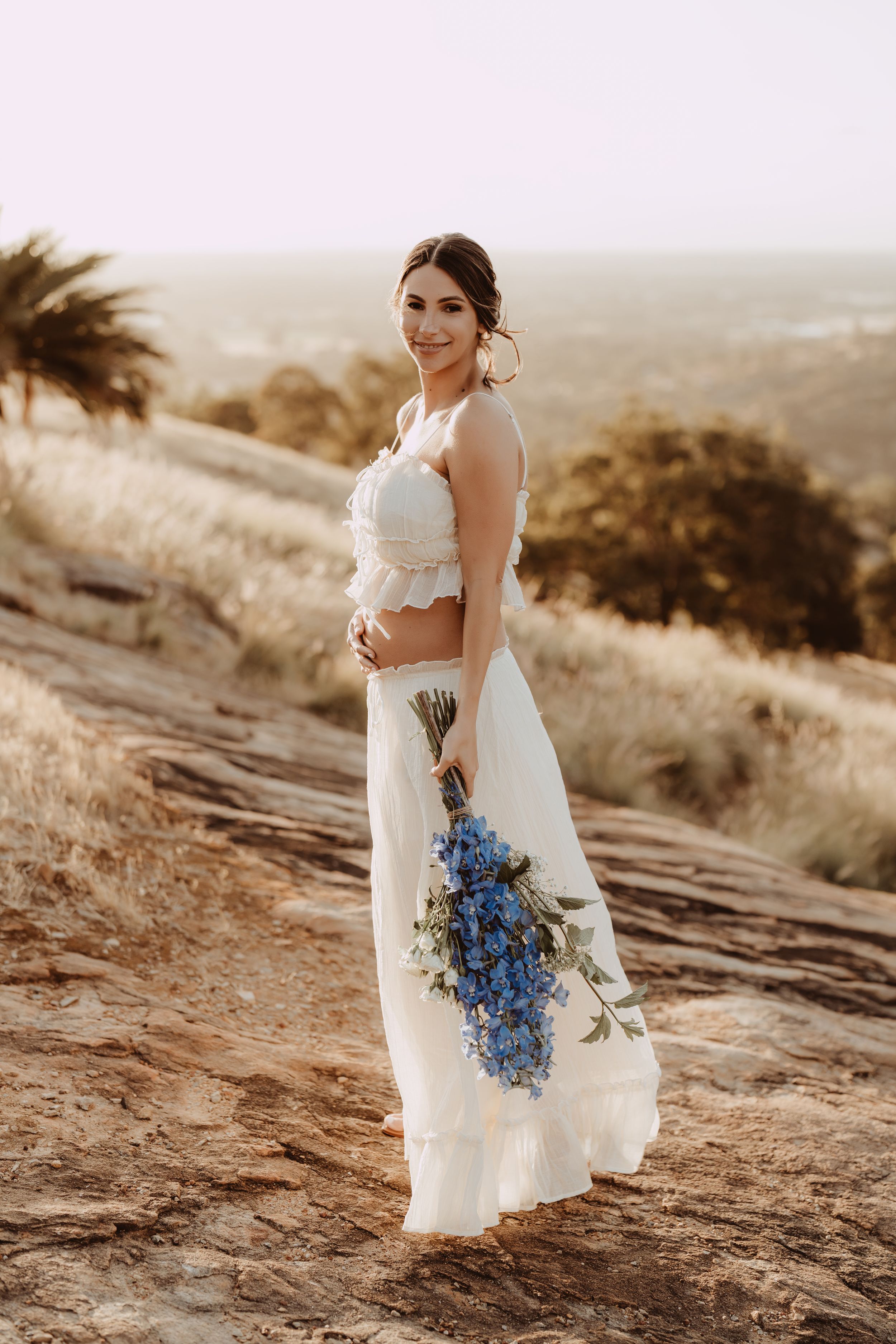 A bride in a white dress holds a vibrant blue bouquet while standing on a scenic desert hillside at sunset.