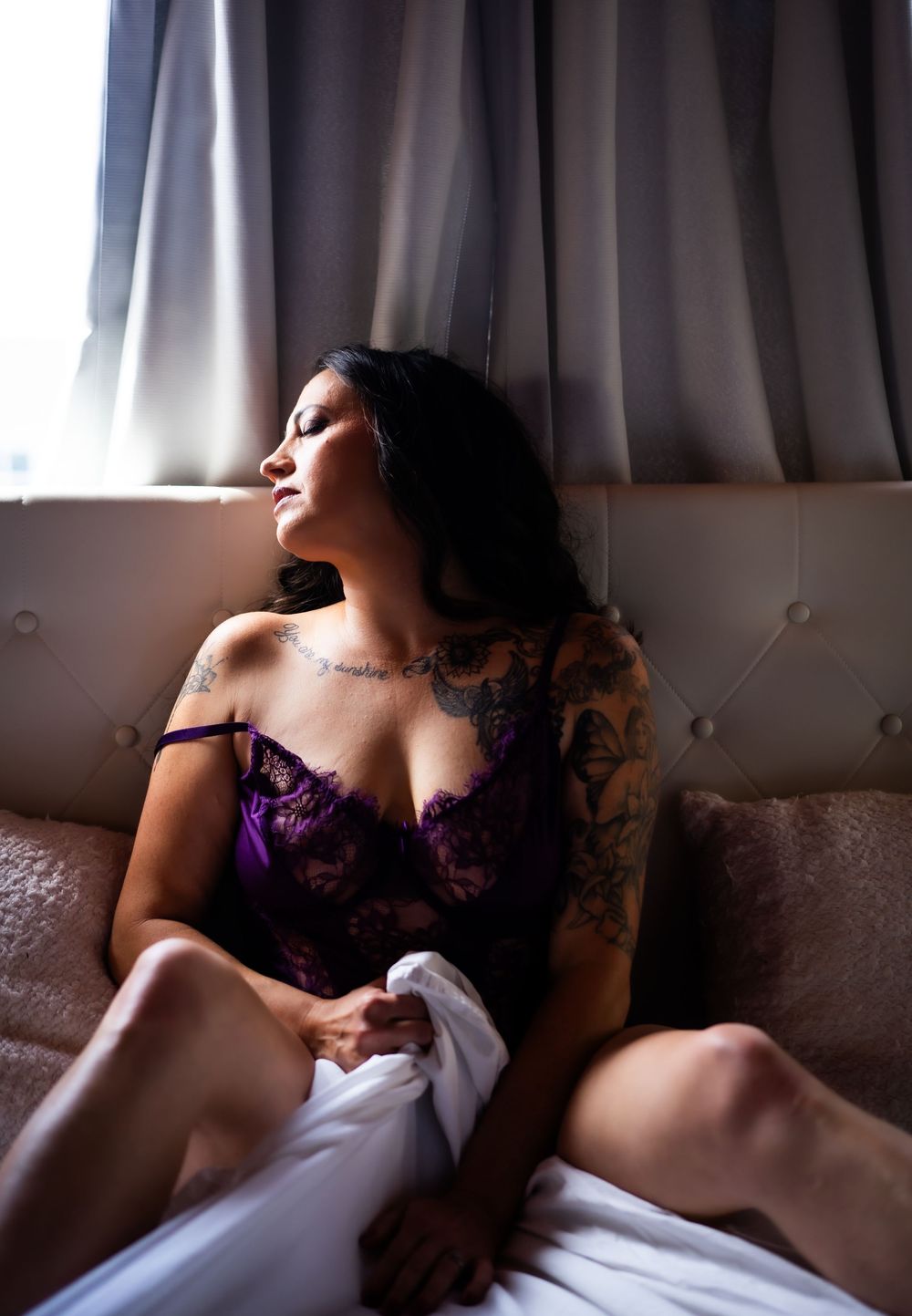 A figure in purple lingerie reclines on a beige couch in soft window light creating an intimate boudoir portrait.