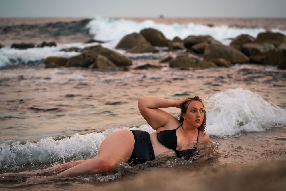 A woman in black swimwear poses on a rocky beach with crashing waves in the background.