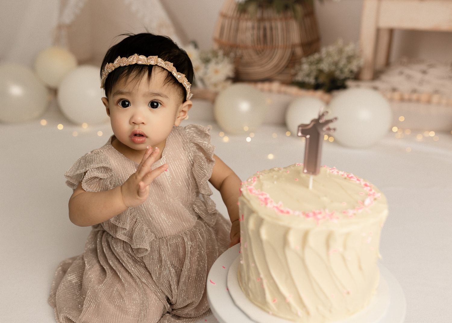 A baby in a beige dress sits near a white birthday cake decorated with pink candles and floral accents.