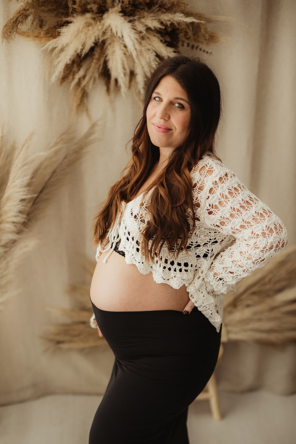 A woman in a black dress with white lace detail poses against a neutral background with dried pampas grass decor.
