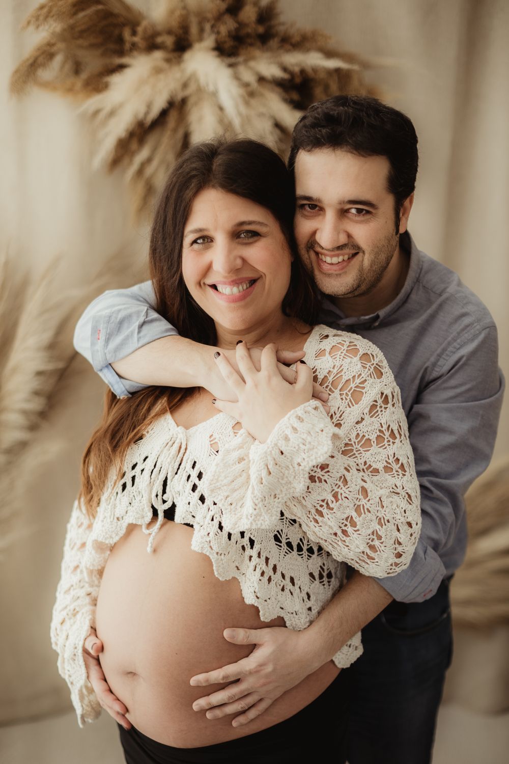 A couple embraces during a maternity photoshoot, with the pregnant woman wearing a white lace top against a neutral background.