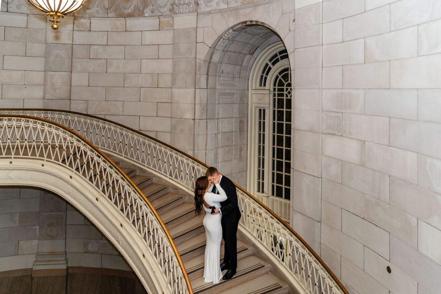 Bride and groom share a romantic embrace on an elegant curved staircase in a grand stone building in Hartford City Hall.