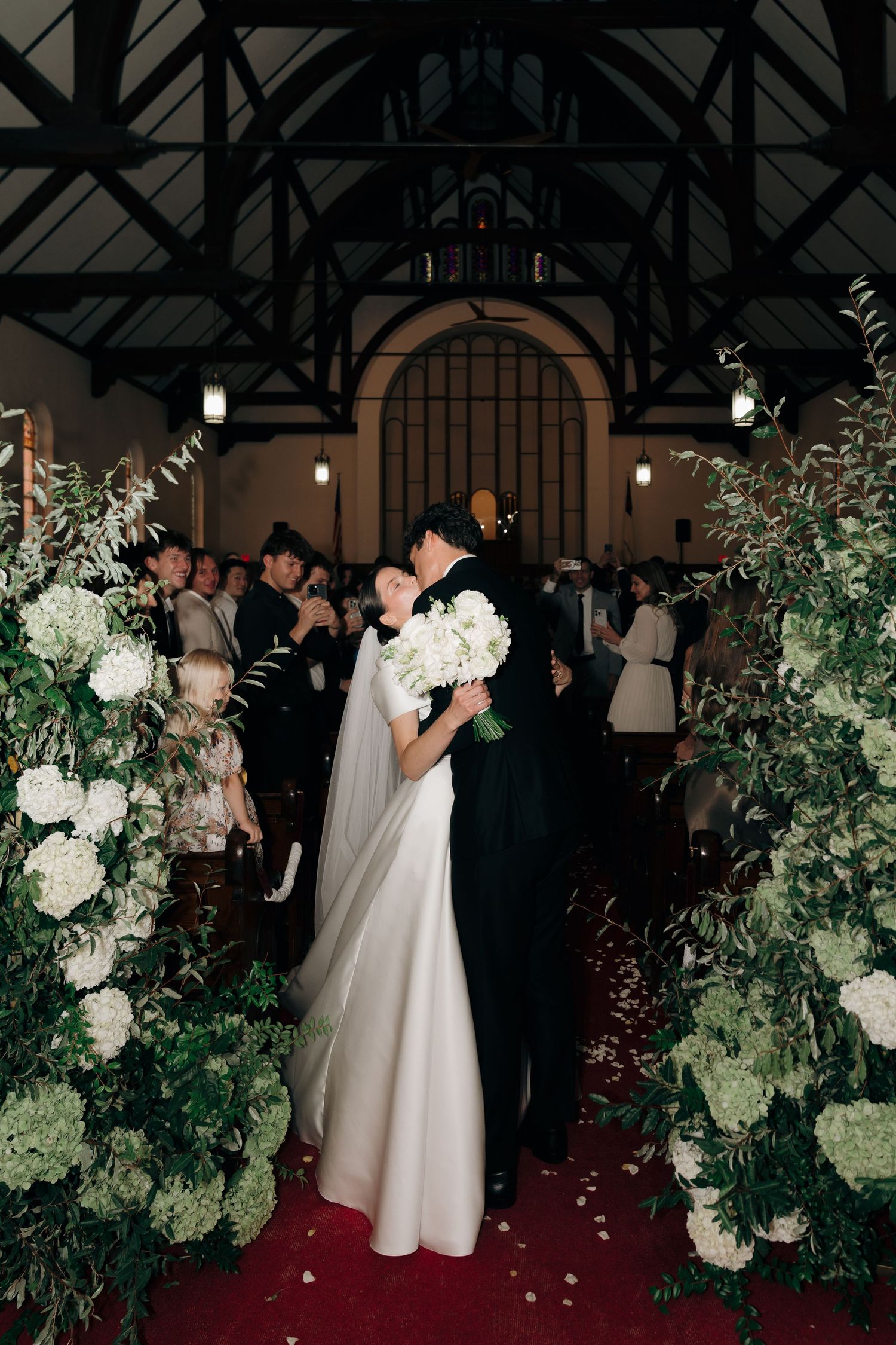 Bride and groom share first kiss at church altar adorned with lush white floral arrangements and greenery.
