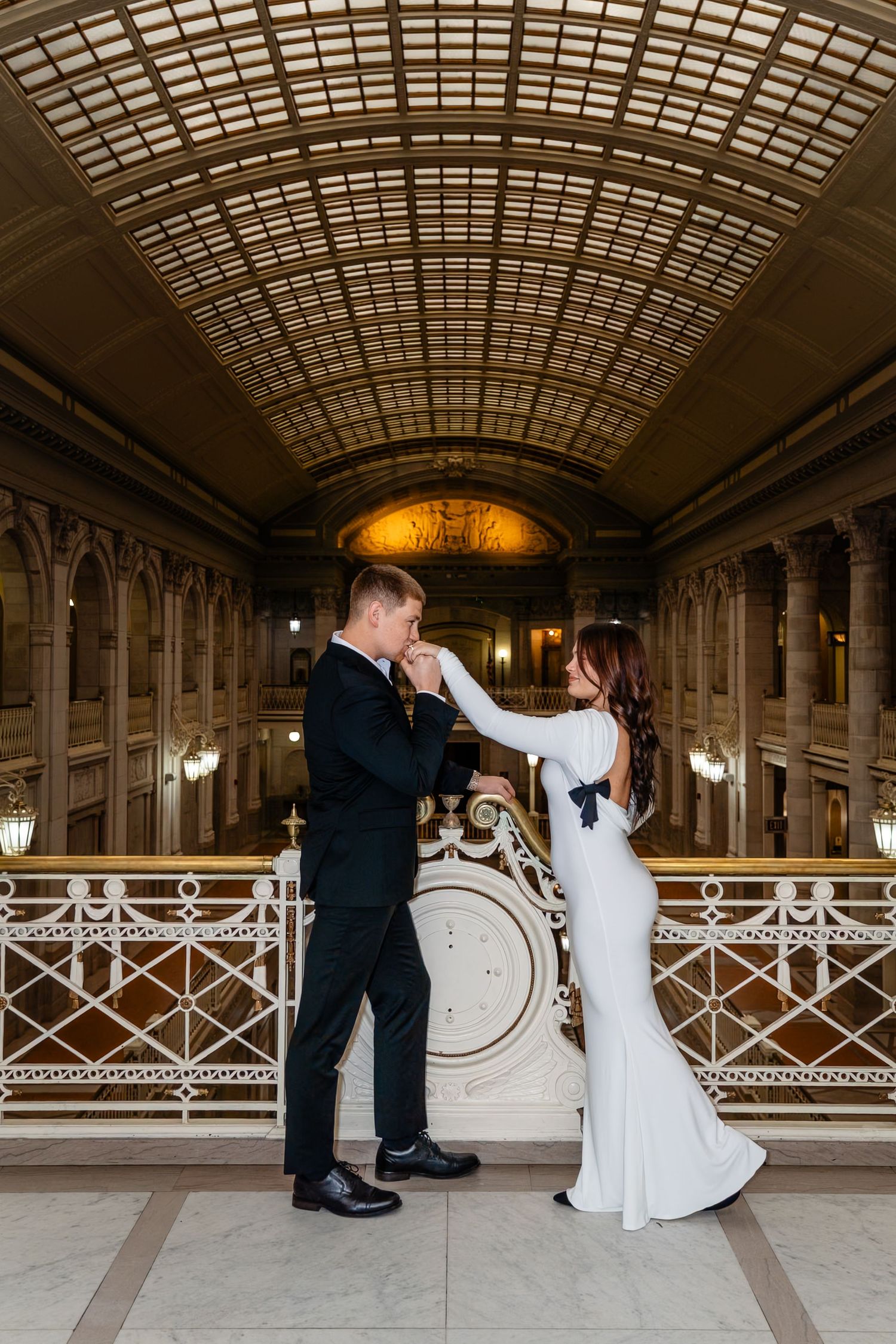 Bride and groom share a romantic hand-kiss moment in an elegant grand hall with arched glass ceiling.