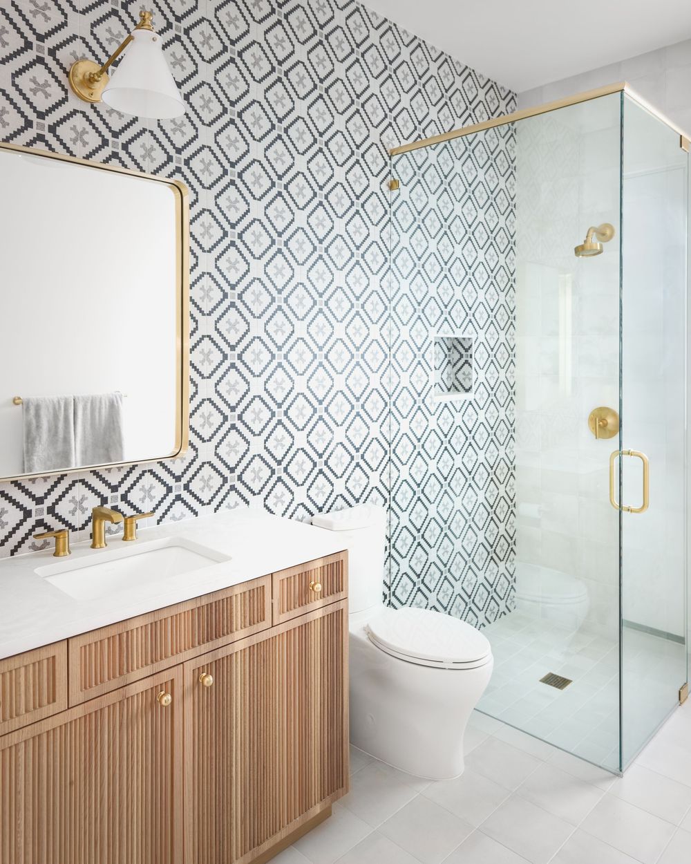 Classic bathroom with diamond patterned wallpaper, wood fluted vanity, gold fixtures, and glass shower.