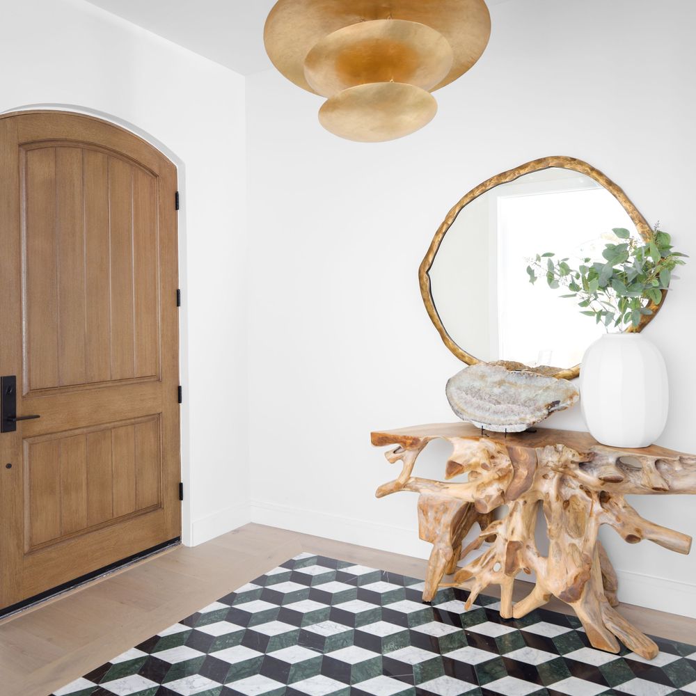 Minimalist entryway with arched wood door, teak root console, geometric floor tiles, and gold mirror.