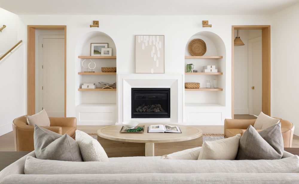 Bright white living room with arched built-in shelves, fireplace, round wood coffee table, and neutral sofa.