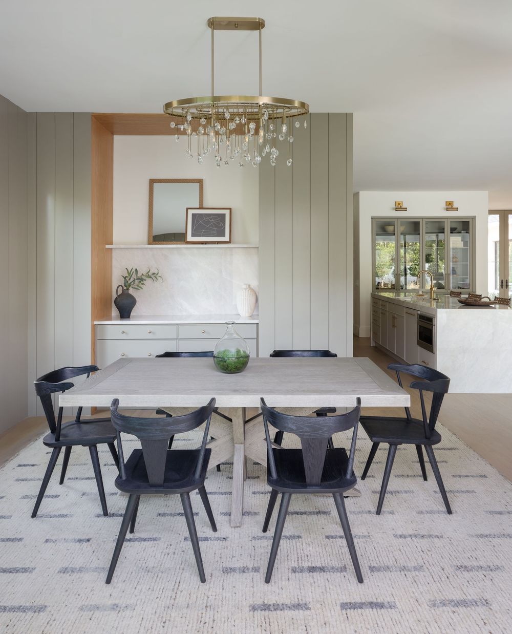 Modern dining room with sage green paneling, light wood table, black chairs, and brass chandelier.