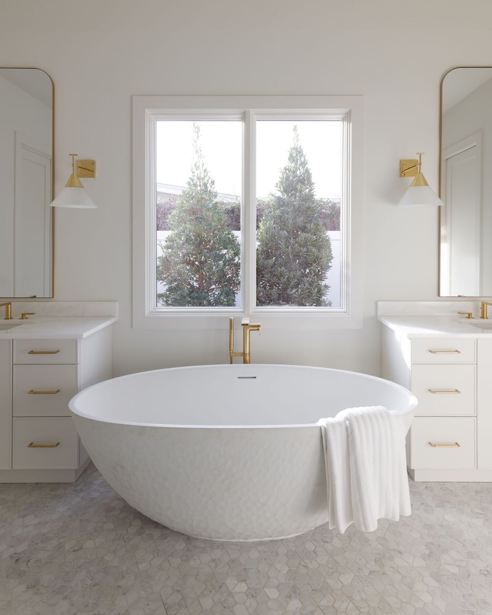 Elegant white bathroom with freestanding oval tub, dual vanities, gold fixtures, and hexagon tile floor.