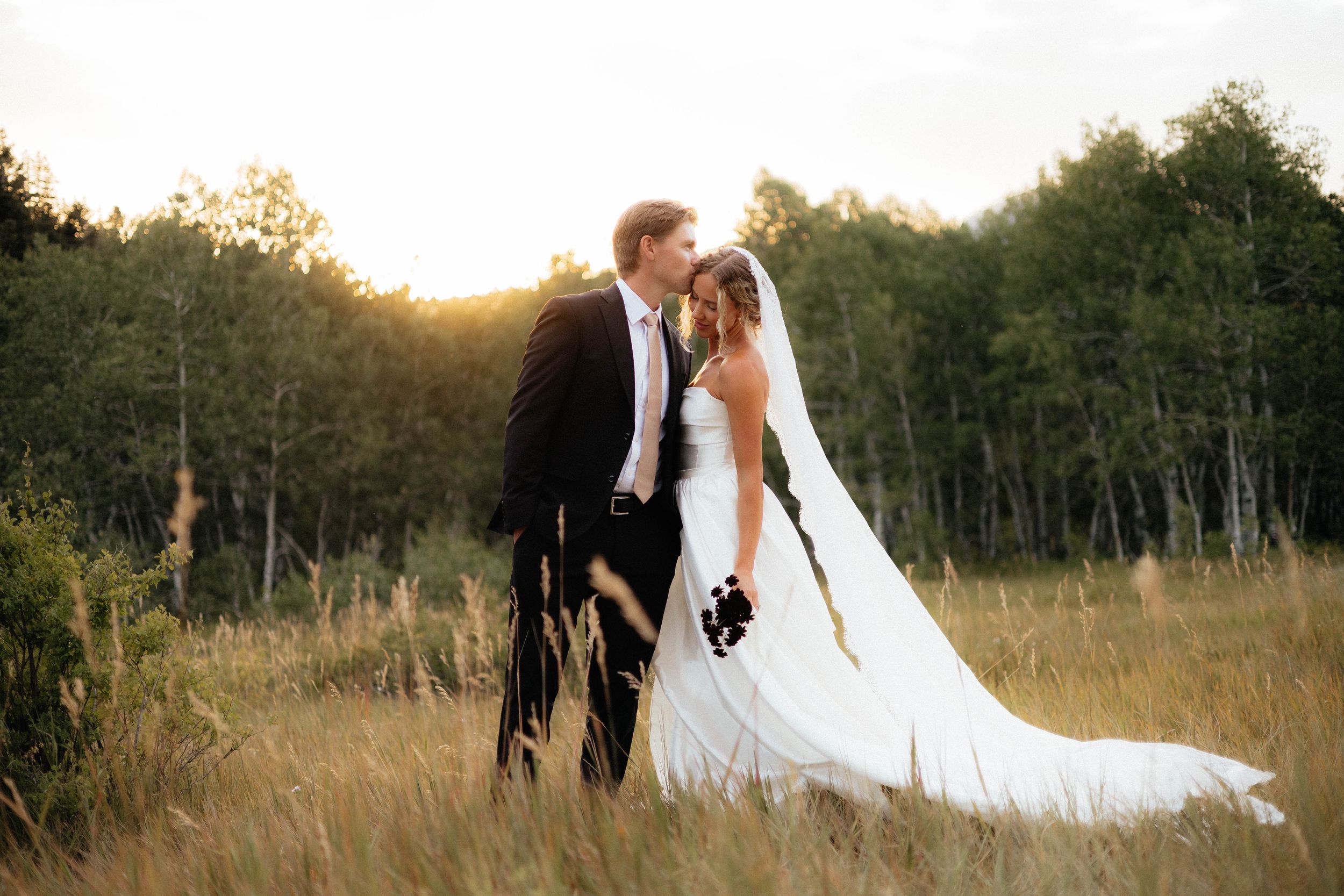 Bride and groom share a romantic moment in a sunlit meadow, surrounded by lush green trees on their wedding day.