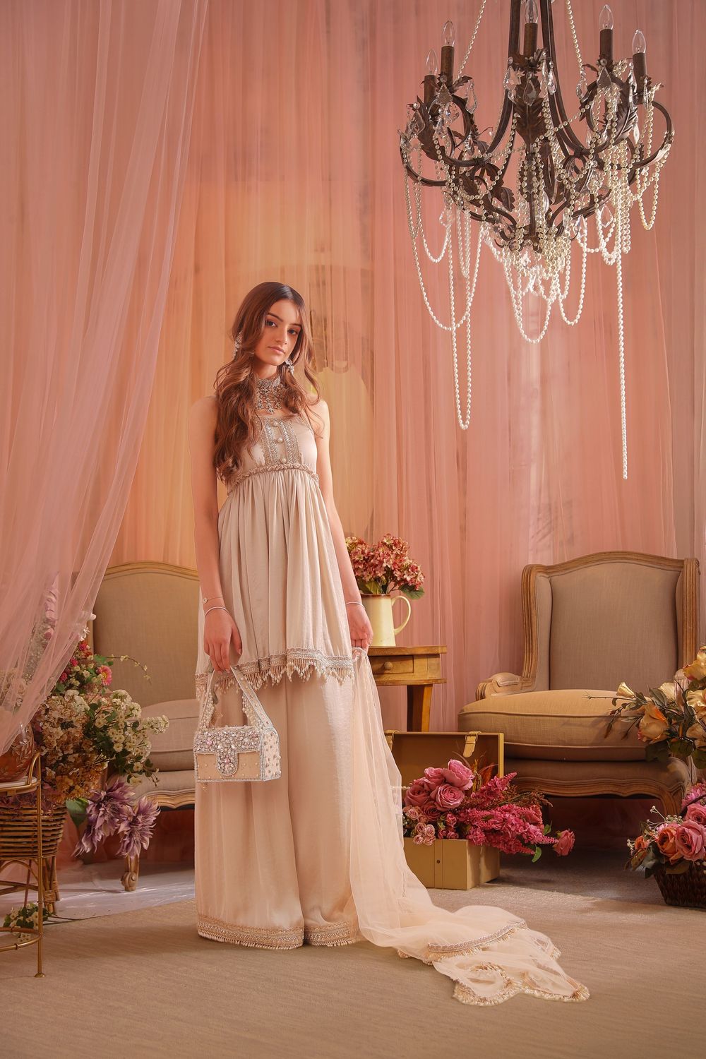 Elegant woman in ivory maxi dress holding crystal bag, standing in romantic pink draped room with chandelier and flowers.