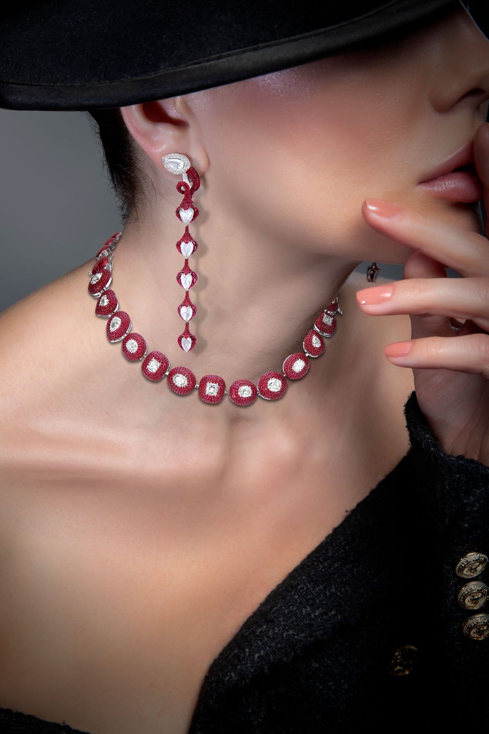 Woman wearing elegant ruby and diamond necklace with matching drop earrings, black hat and jacket, manicured nails.