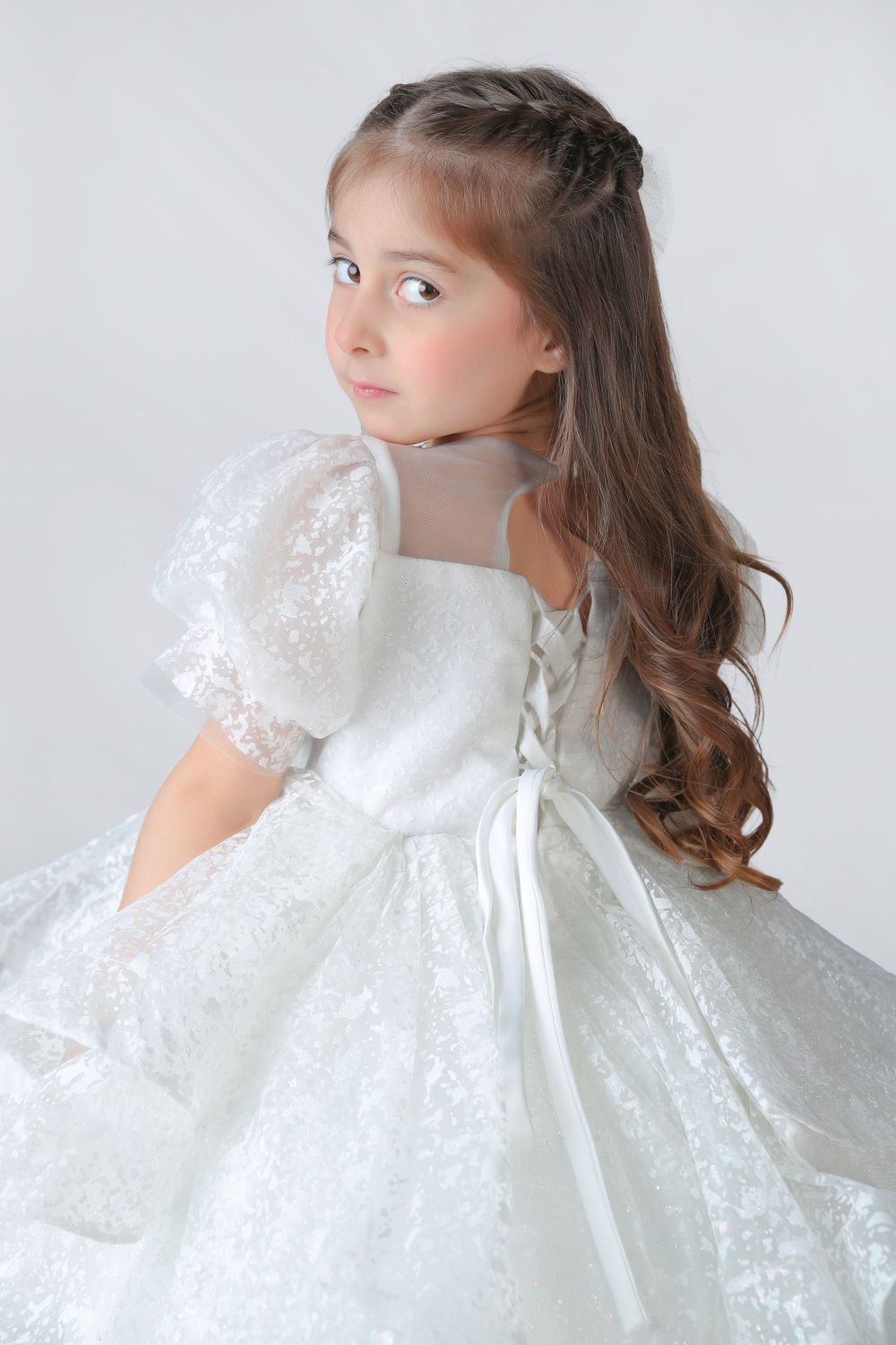 Young girl in white lace dress with puff sleeves and long curly hair, looking over shoulder gracefully.