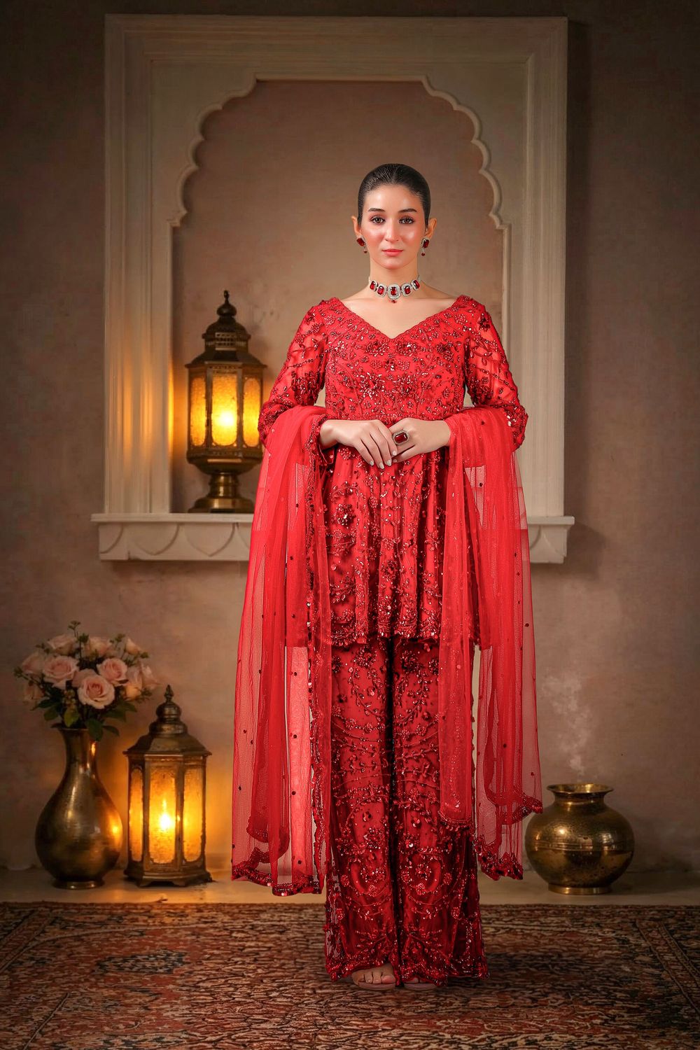 Woman in red embroidered Pakistani sharara suit with dupatta, standing in elegant traditional setting with lanterns.