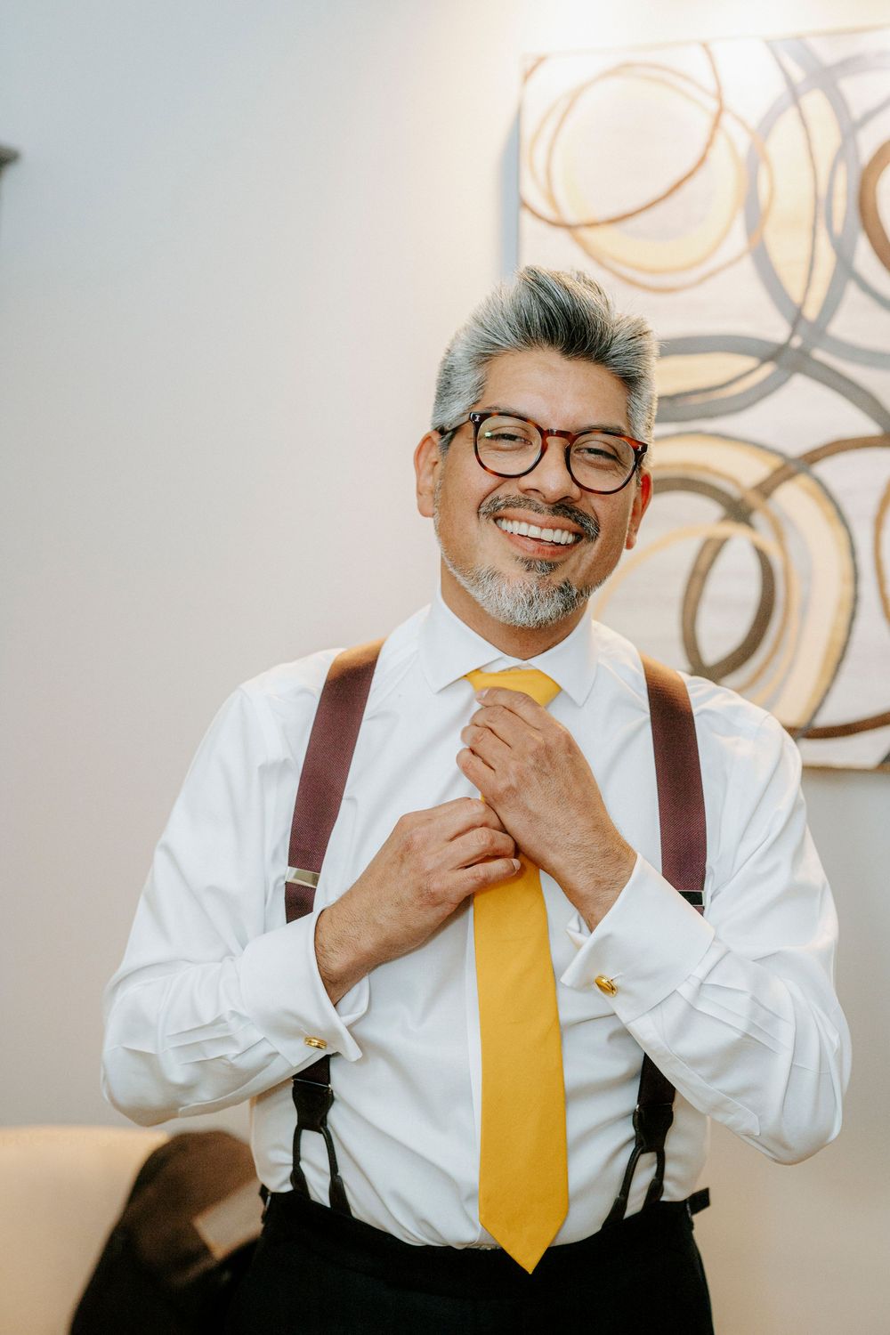 Smiling middle-aged groom adjusting yellow tie with suspenders and white shirt, wearing glasses.