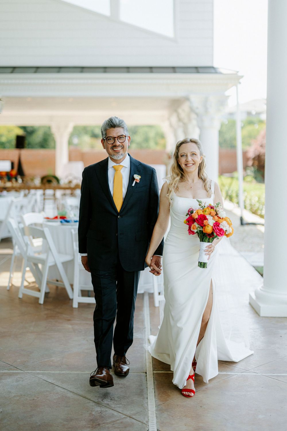 Bride and groom walk hand-in-hand at outdoor wedding venue, she holds colorful bouquet wearing white gown with red heels.