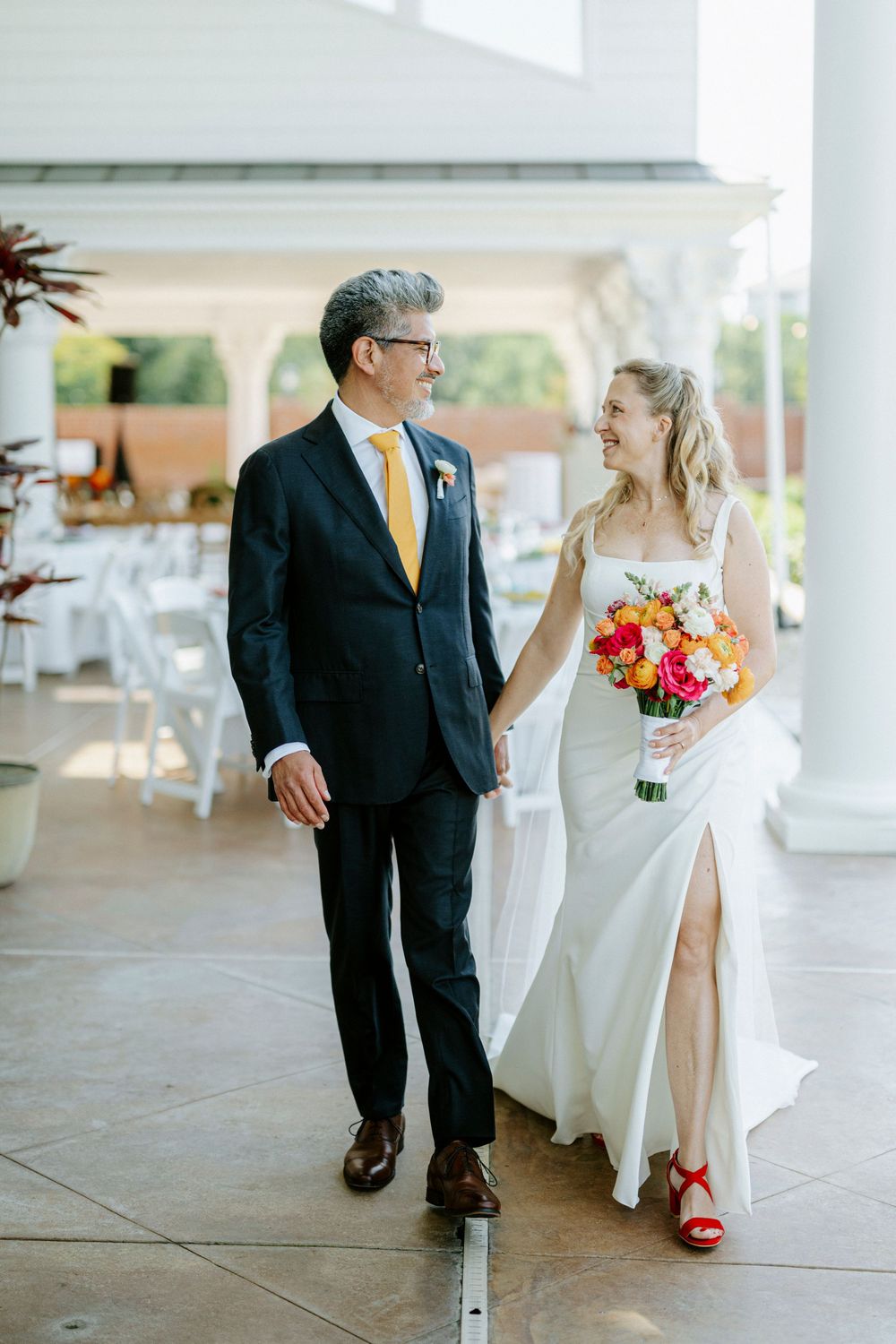 Bride in white gown with red heels and groom in navy suit with yellow tie walk hand in hand at wedding venue.