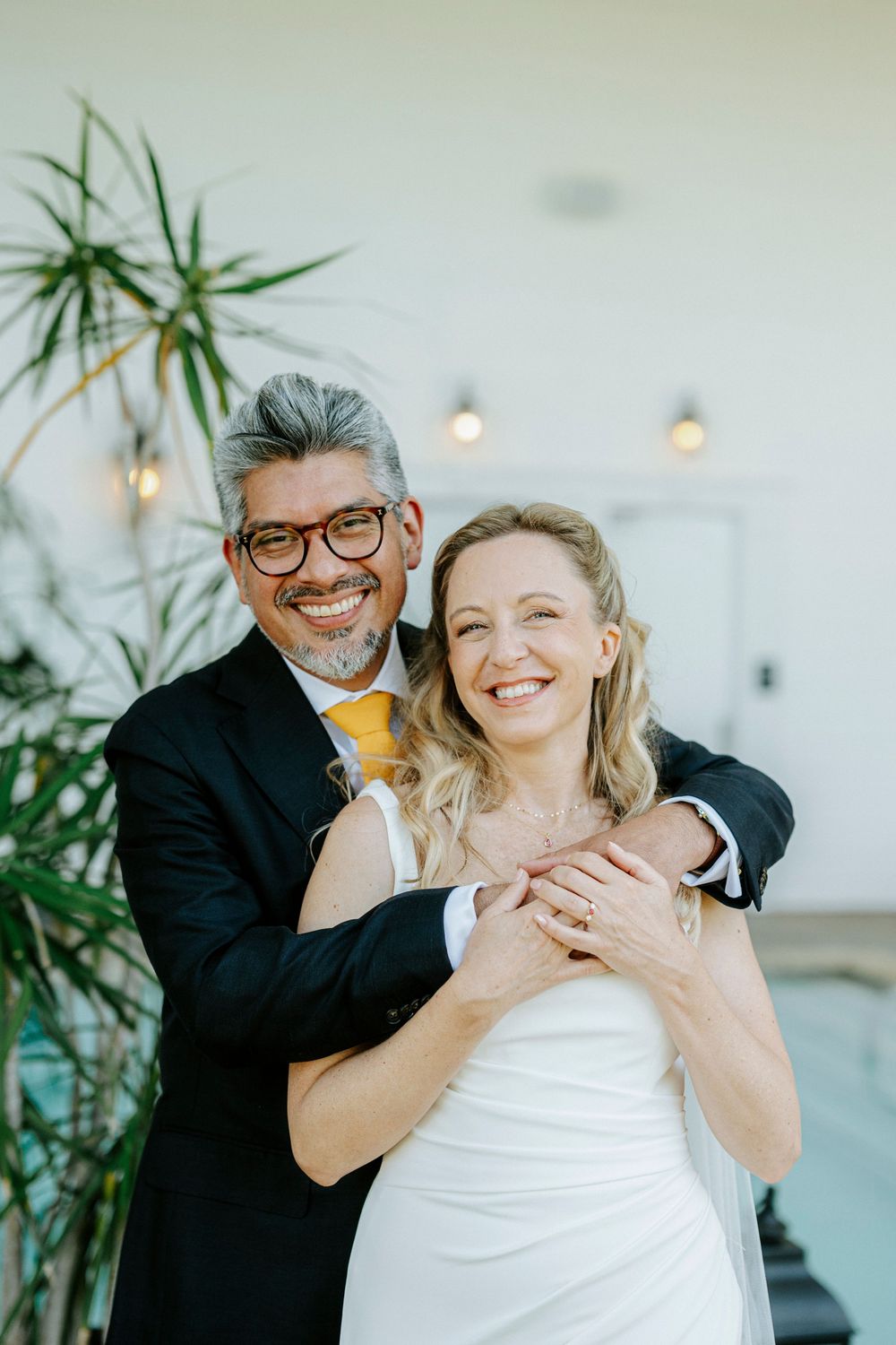 A smiling couple in wedding attire embraces indoors, groom in dark suit with yellow tie, bride in white dress.