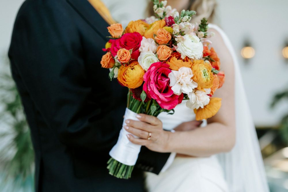 Bride holding vibrant wedding bouquet with orange, pink, and white ranunculus flowers while standing with groom.