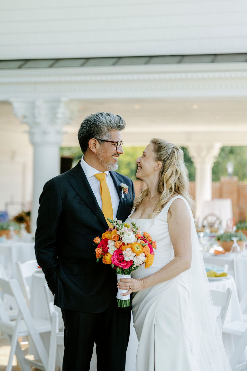 Bride and groom share a loving glance as she holds a vibrant bouquet of orange and pink flowers outdoors.