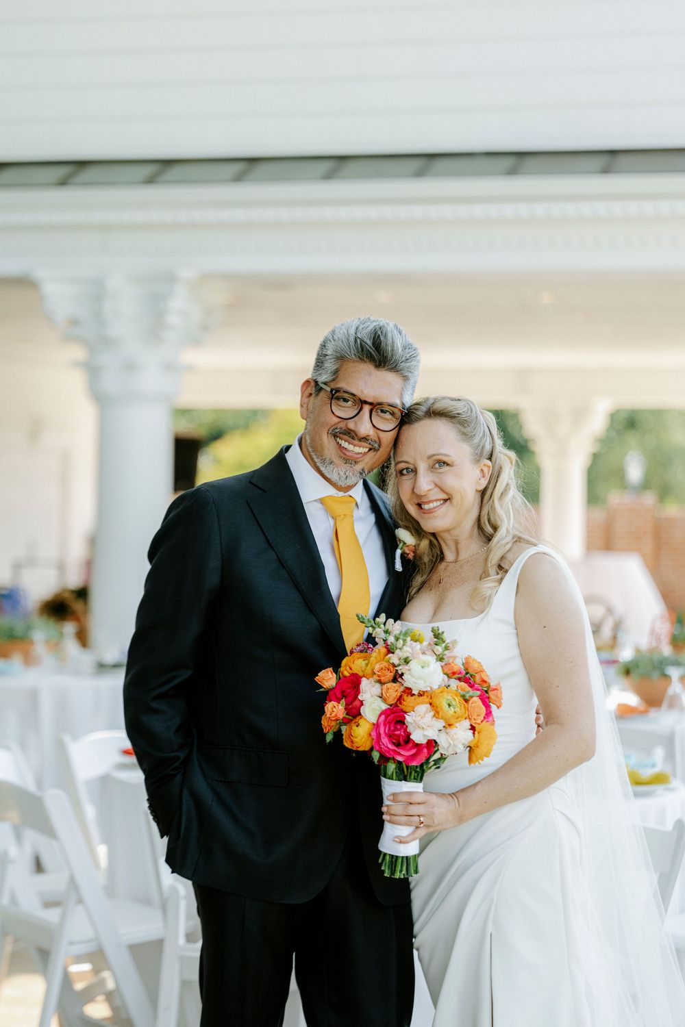 Smiling newlyweds pose together at outdoor wedding, groom in dark suit with yellow tie, bride holding vibrant floral bouquet.