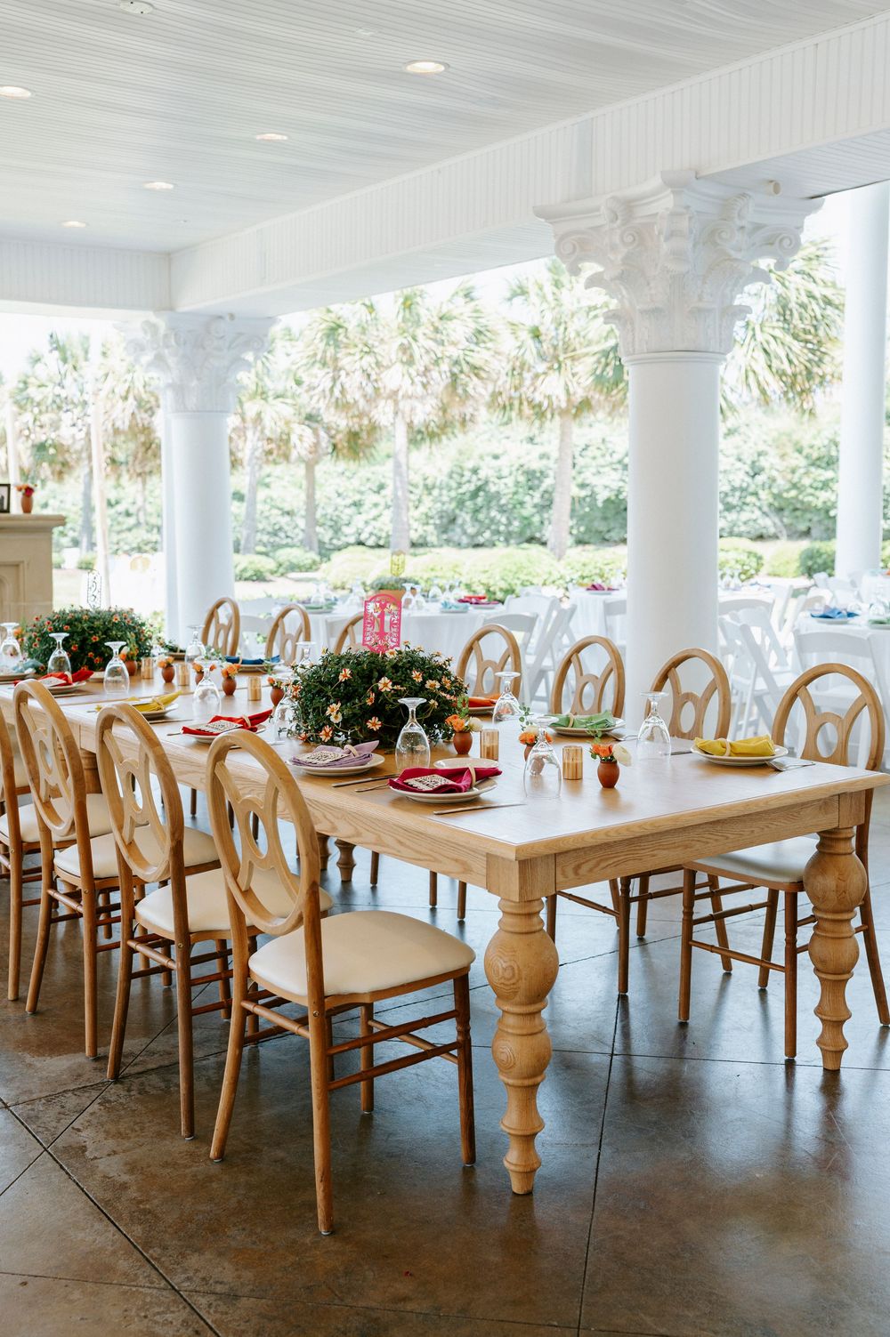 Elegant wedding reception table with natural wood chairs, floral centerpiece, and red accents on covered porch.