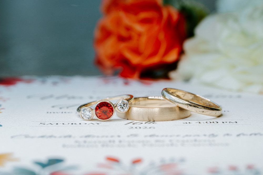 Three gold wedding rings with diamond and ruby gemstones displayed on paper with an orange rose in background.