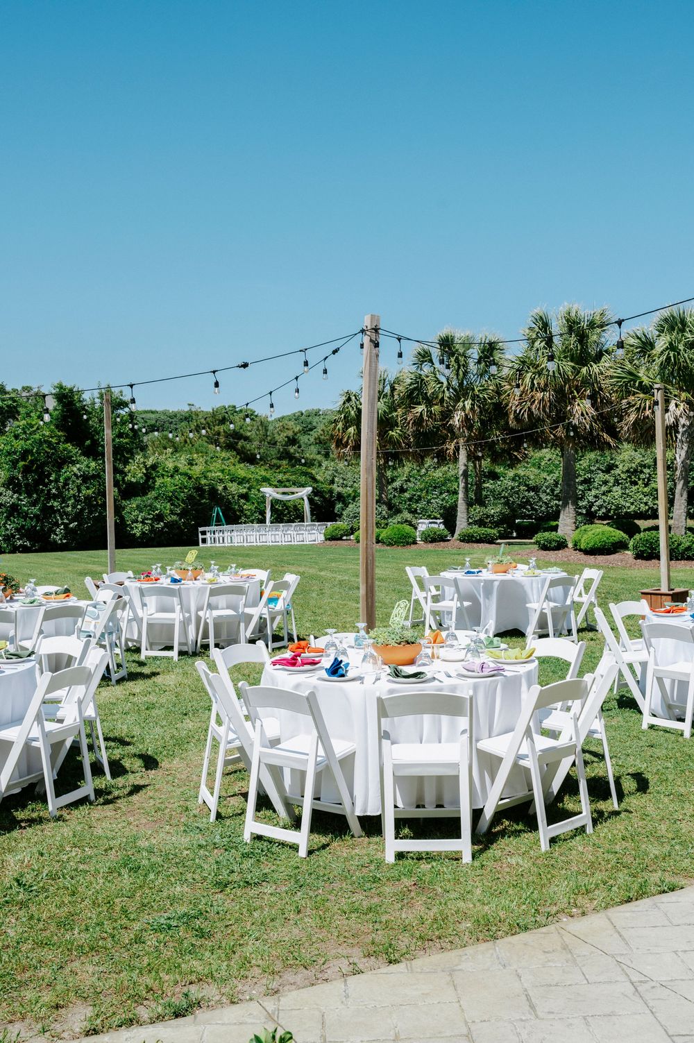 Outdoor wedding reception with round white-clothed tables, white folding chairs, string lights, and lush palm tree backdrop.