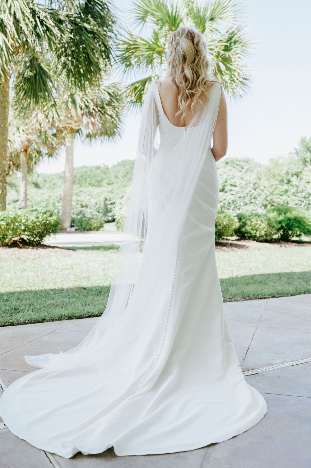 Bride in elegant white fitted wedding gown with long veil and train stands in lush tropical garden setting.