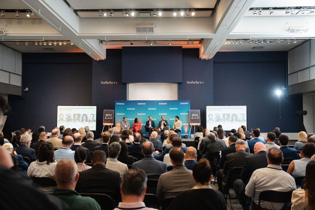 Large conference audience listens to panel of speakers on a blue-lit stage at a professional business event.