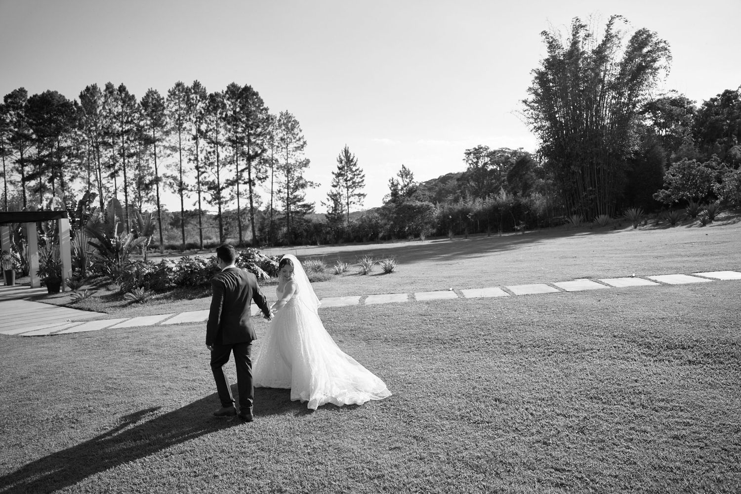 Black and white outdoor wedding ceremony with bride in veil walking toward guests gathered on lush garden lawn.
