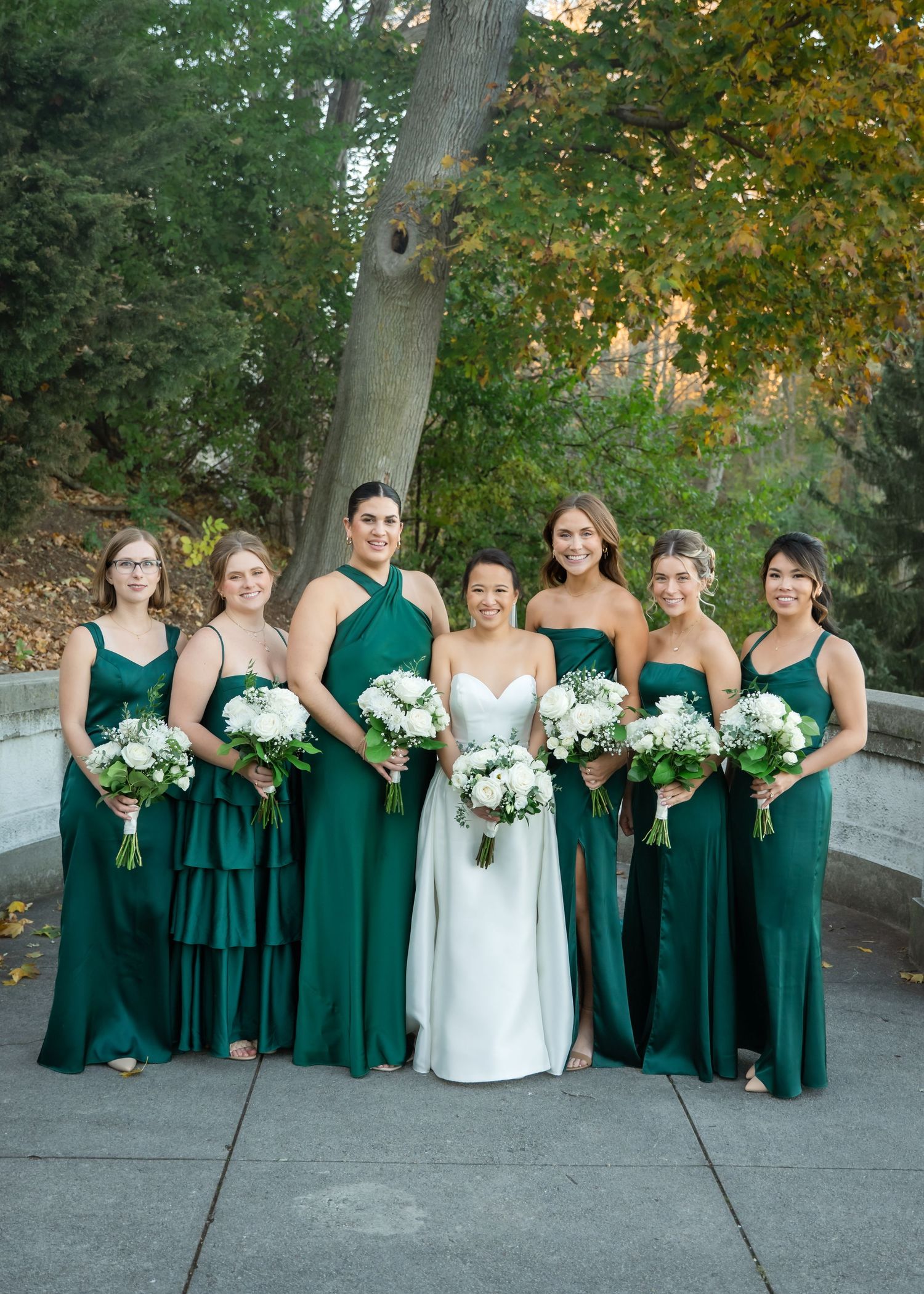 Bride in white gown with six bridesmaids in emerald green dresses holding white bouquets outdoors.