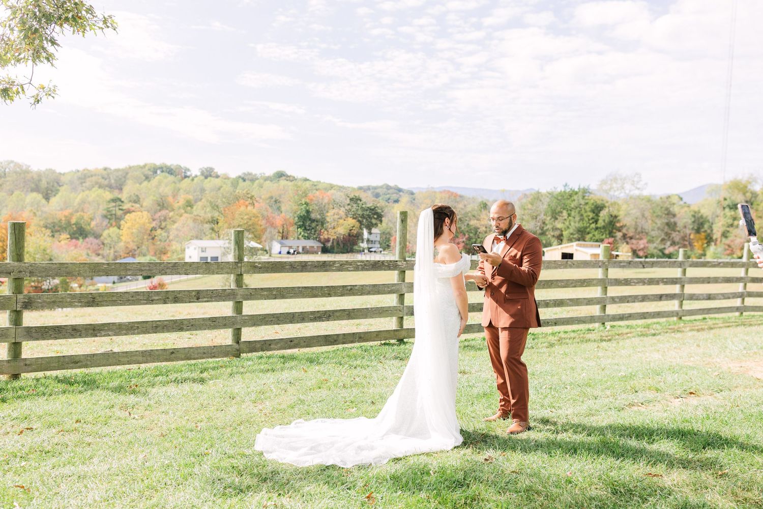 Bride and groom share a first look moment on a green lawn with a wooden fence and autumn trees backdrop.
