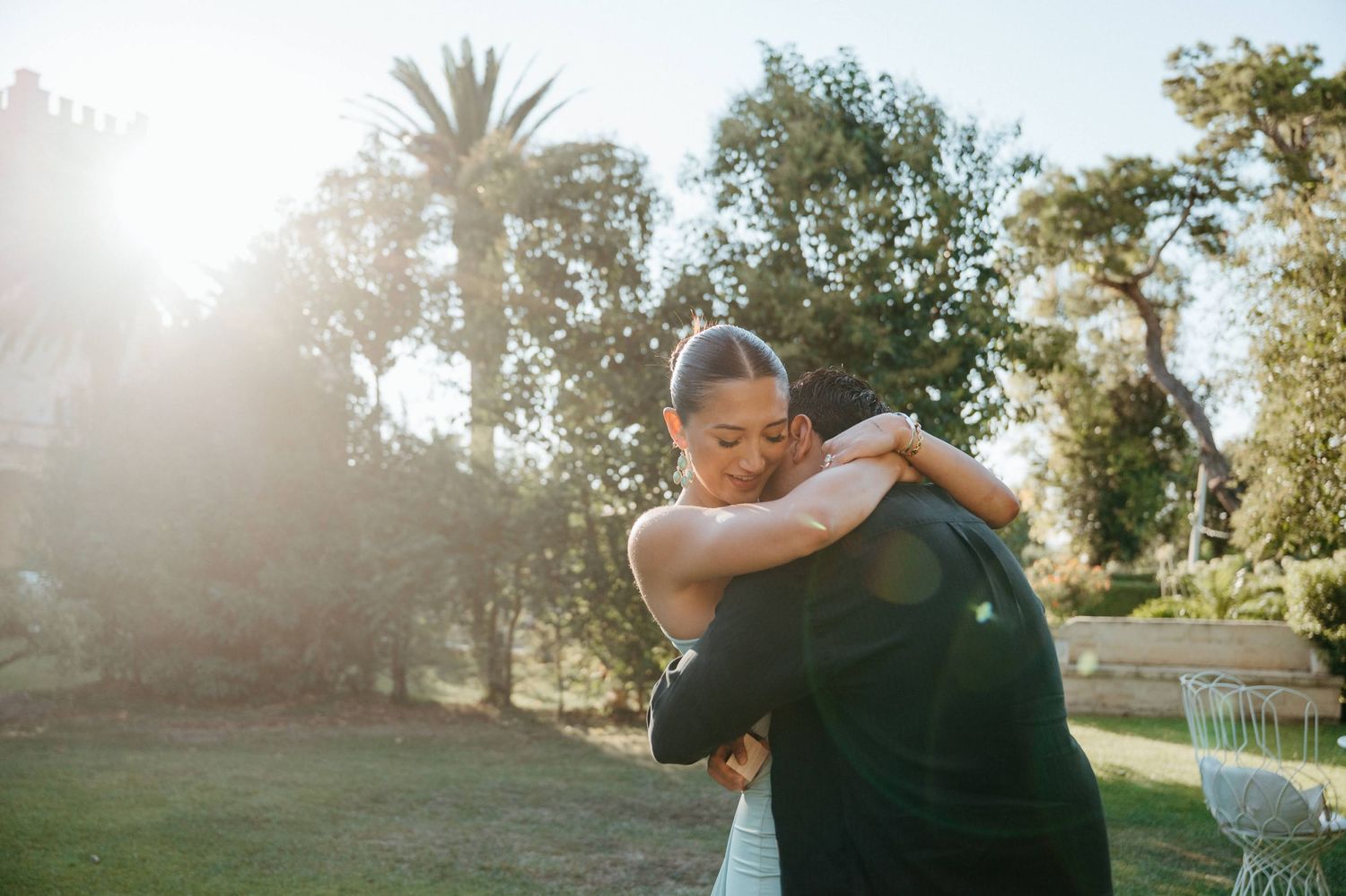 A couple embraces tenderly in a golden field, woman hugging man from behind among wildflowers.