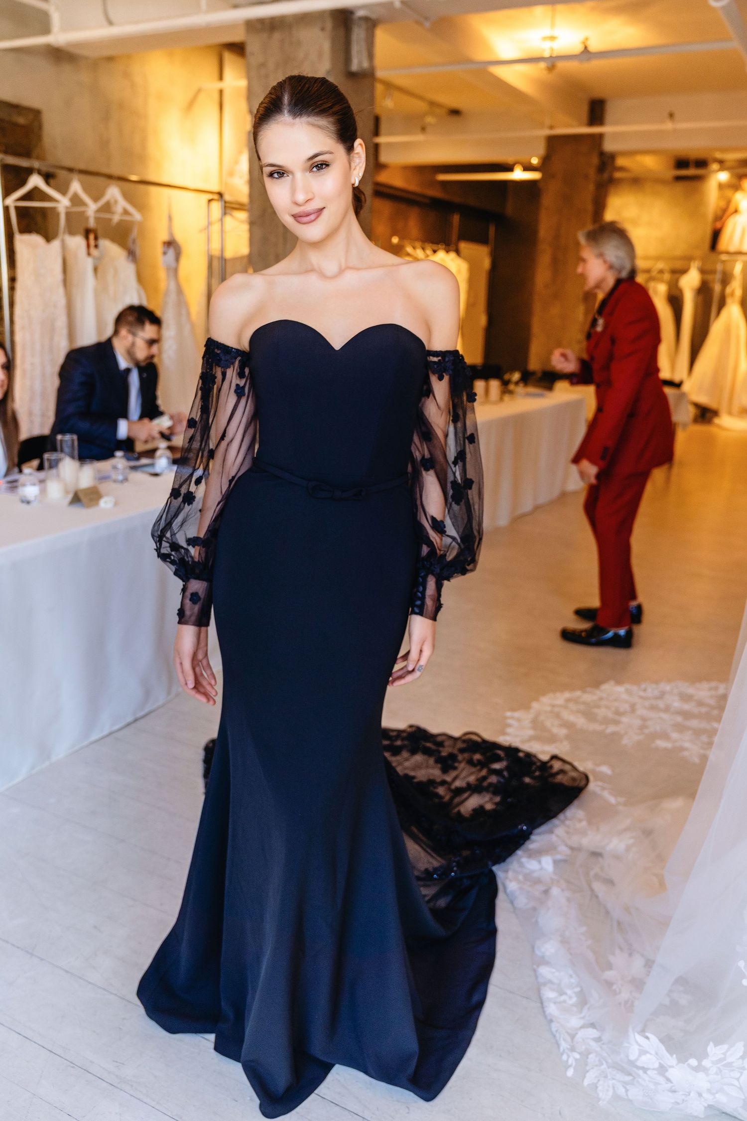 Woman in elegant navy off-shoulder gown with sheer lace sleeves poses in a bridal boutique.