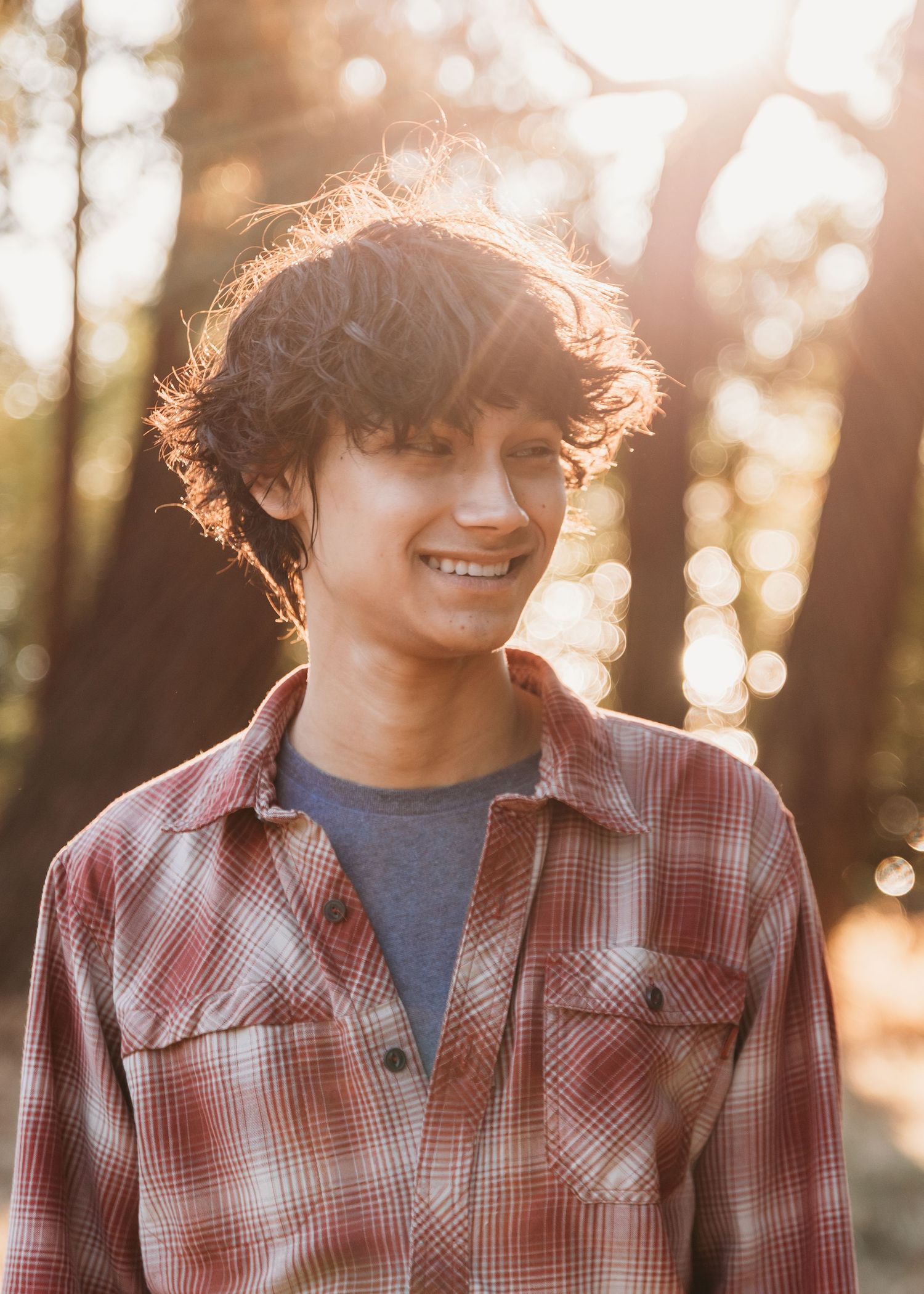 Smiling young man with curly hair wearing a plaid flannel shirt outdoors in golden sunlight among trees.