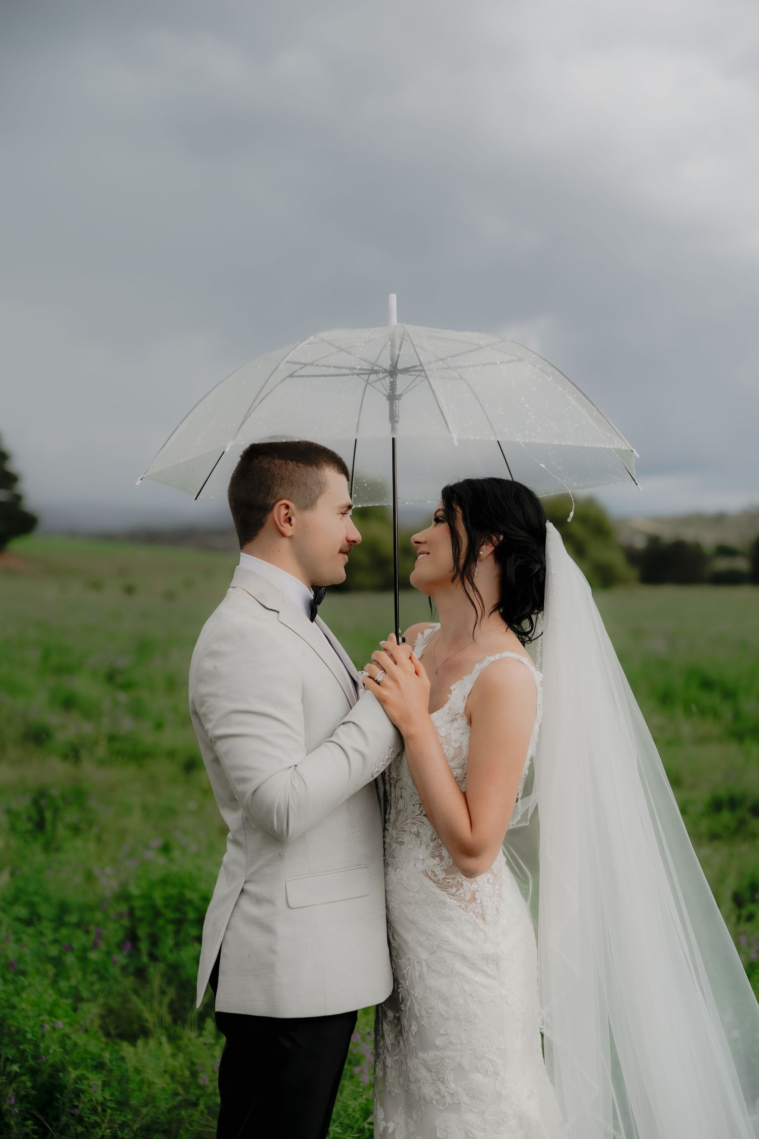 A bride and groom share a romantic moment under a clear umbrella in a green field on a cloudy wedding day.